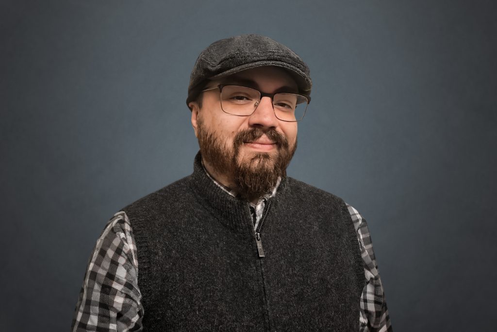 headshot of Joseph Miranda wearing newsboy cap and glasses.